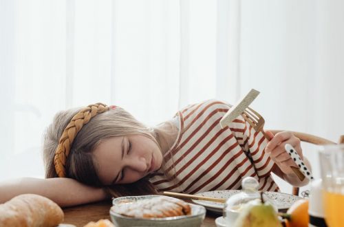 A girl in a striped shirt resting her head on a table surrounded by breakfast foods, holding a fork.