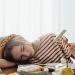A girl in a striped shirt resting her head on a table surrounded by breakfast foods, holding a fork.